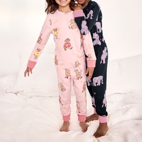 Two children wearing pink bear-print and navy elephant-print long-sleeve pyjama sets, standing on a white bed.