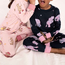 Two children wearing matching pink and navy dinosaur and bear print pyjama sets, seated together on a white bed.