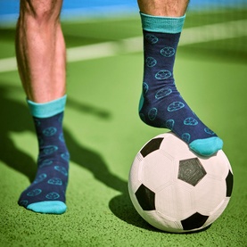 Model wearing Gentlemen's Hardware Football Socks Tin, navy blue socks with teal football print and teal cuffs, foot resting on football