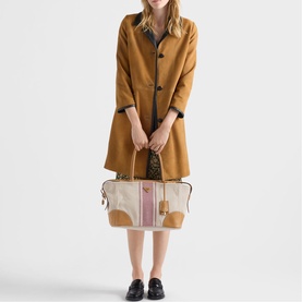 Model carrying Prada Accessories neutral canvas tote bag with tan leather trim and pink stripe detail, styled with a camel coat and black loafers