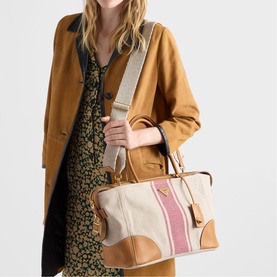 Model carrying Prada Accessories neutral canvas and tan leather top-handle bag with pink stripe detail, gold hardware, and wide fabric crossbody strap