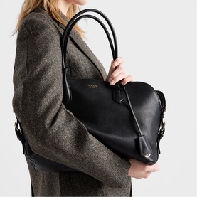 Model carrying Prada Accessories black leather tote bag with gold-tone hardware and rolled top handles, styled with a brown tweed blazer