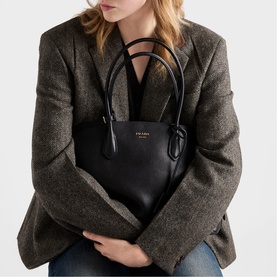Model carrying Prada black leather tote bag with gold-tone logo and dual top handles, styled with a grey herringbone blazer