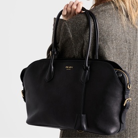 Model carrying Prada Accessories black leather tote bag with gold-tone hardware and short top handles, styled with a grey tweed jacket.