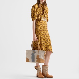 Model carrying Prada Accessories neutral tote bag with stripe detail and tan leather trim, styled with mustard floral dress and tan ankle boots