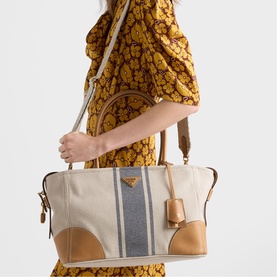 Model carrying Prada Accessories neutral canvas tote bag with tan leather trim, grey stripe detail, gold-tone hardware and adjustable shoulder strap