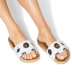 Model wearing Kurt Geiger London white slides with football print strap and gold flatform sole, styled with white leggings