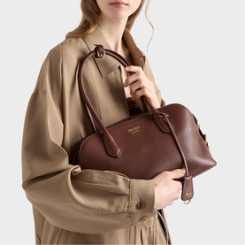 Model carrying Prada Accessories brown leather structured top-handle bag with gold hardware and detachable shoulder strap, styled with beige coat