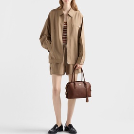 Model carrying Prada Accessories brown leather top-handle bag with gold-tone hardware, styled with beige oversized shirt, shorts and black loafers.