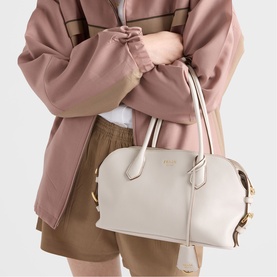 Model carrying a cream structured dome bag with gold hardware and top handles, styled with a pink oversized jacket and tan shorts