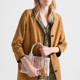 Model carrying a small striped canvas and leather tote bag with gold triangle logo, styled with a camel coat and floral dress.