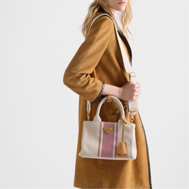 Model carrying a structured canvas tote bag in cream with pink stripe detail, gold-tone hardware and tassel, styled with a camel coat