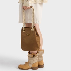 Model carrying Prada Accessories brown mini bucket bag with rope handles and gold triangle logo, styled with camel boots and cream skirt.