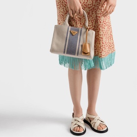 Model carrying Prada Accessories neutral canvas tote bag with blue stripe, gold-tone hardware and wooden top handles, styled with fringed dress and