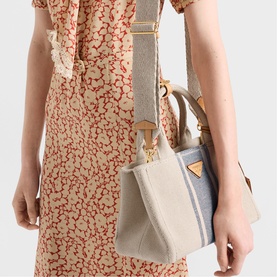Model carrying Prada Accessories neutral canvas mini bucket bag with gold hardware and wide fabric crossbody strap, styled with floral dress