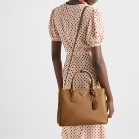 Model carrying Prada Accessories brown saffiano leather tote bag with gold hardware, tassel detail, and crossbody strap, styled with polka dot dress.