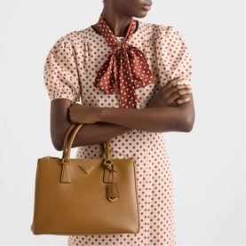 Model carrying Prada Accessories brown structured tote bag with gold-tone hardware and short top handles, styled with a polka dot dress and neck scarf