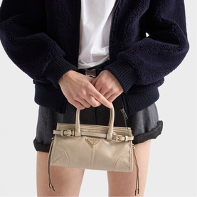 Model carrying Prada Accessories neutral leather top-handle bag with gold-tone triangle logo, styled with dark shorts and navy fluffy jacket.