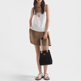 Model carrying Prada Accessories black bucket bag with gold hardware, styled with white tank, tan shorts and white sandals