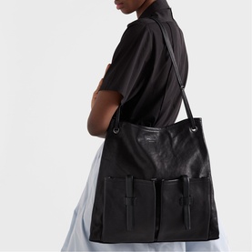 Model carrying Prada Accessories black leather tote bag with front strap detail, worn on shoulder over white skirt outfit.