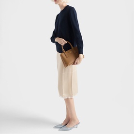 Model carrying a tan structured tote bag with gold hardware, styled with navy top, cream midi skirt and grey heels.