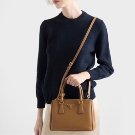 Model carrying a tan Prada structured tote with gold-tone hardware and adjustable crossbody strap, styled with navy knit and cream trousers