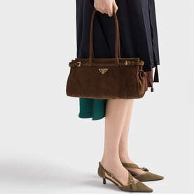 Model carrying Prada Accessories brown suede tote bag with gold logo hardware and top handles, styled with green skirt and kitten heels.