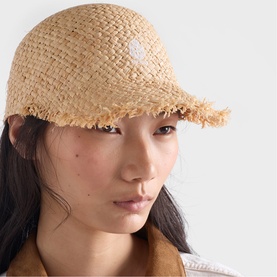 Model wearing a natural raffia baseball cap with frayed brim edge and small logo patch, styled with an orange jacket