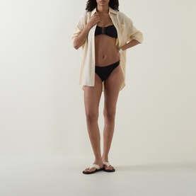 Model wearing Sea Level black bikini bottom styled with a cream oversized shirt and tan slide sandals.