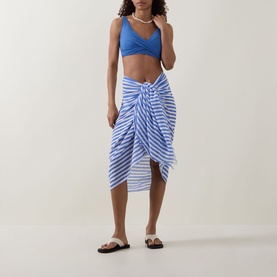 Model wearing Sea Level Essentials V-Front Bikini Bottoms in Denim Blue, styled with matching blue bikini top, striped sarong and white sandals.