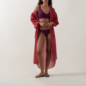 Model wearing Sea Level Essentials Cross Front Multifit Bikini Top in Claret with matching bottoms and a red striped open cover-up and flat sandals.