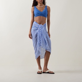 Model wearing Sea Level Essentials Cross Front Tie Back Bikini Top in Denim Blue with a blue striped sarong skirt and white sandals.