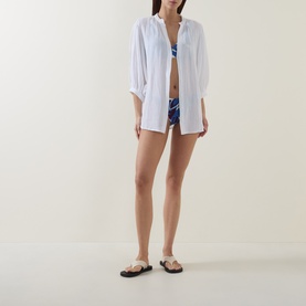 Model wearing Sea Level white cotton cover-up shirt, styled with blue printed bikini bottoms and black toe-post sandals.