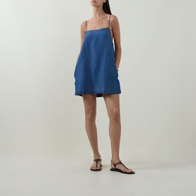 Model wearing Sea Level Denim Surf Mini Shift Dress in denim, straight neckline, narrow straps, flared skirt; styled with brown flat sandals