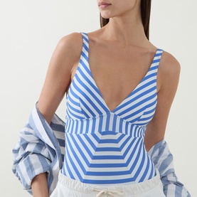 Model wearing Sea Level denim blue stripe V-neck swimsuit with geometric panel detail, styled with striped shirt and white shorts