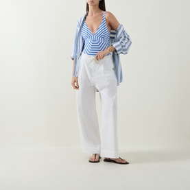Model wearing Sea Level denim blue stripe swimsuit with white wide-leg trousers, blue open shirt and tan flat sandals.