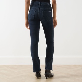Model wearing Seven Jeans Tailorless Mid-Rise Bootcut Jeans in dark blue, rear view showing logo badge and split unfinished hems, styled with black