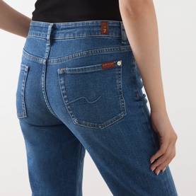 Model wearing Seven Jeans blue bootcut jeans, close-up of back pocket detail with signature logo patch, paired with black top.