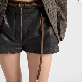 Model wearing black textured leather-look shorts with zip front and brown leather belt, styled with a grey herringbone blazer