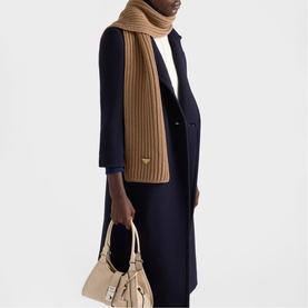 Model wearing a camel ribbed knit scarf with navy coat, carrying a beige structured bag with gold hardware.