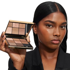 Model with open contour and highlight palette in warm nude tones, gold-edged compact held open to camera, wearing black blazer.