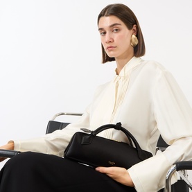 Model carrying Alaia Le Teckel Clutch Bag in Noir silk satin, gold-tone hardware, tubular handles, styled with cream blouse and black trousers