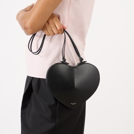 Model carrying Alaia Le Coeur Crossbody Bag in Noir; heart-shaped black leather with silver-tone hardware and top handles, styled with pink top.