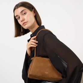Model carrying Alaia Le Bouledogue Suede Shoulder Bag in Terre D'Ombre with gold-tone hardware and tubular strap, styled with black sheer top
