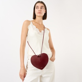 Model carrying Alaia Le Coeur Crossbody Bag in Rouge Grenat, heart-shaped red leather with silver-tone hardware, styled with white lace cami and