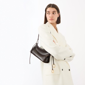 Model carrying Alaia Le Click Small Shoulder Bag in Marron Fonce, gold-tone hardware, adjustable strap, styled with cream coat