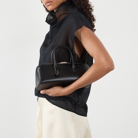 Model carrying Ala&iuml;a Delta Small Tote Bag in noir nubuck calfskin, gold-tone hardware and branded tie detail, styled with black sleeveless top.
