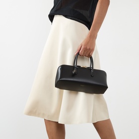 Model carrying Alaia Delta Small Tote Bag in Noir nubuck calfskin, gold-tone hardware, top handles, styled with cream midi skirt and black top