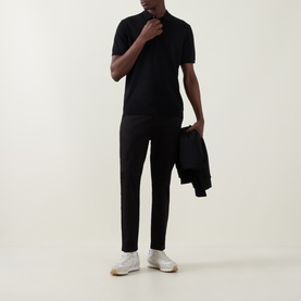 Model wearing Arne Tailored Slim-Leg Chinos in black, styled with a black polo shirt and white trainers, holding a jacket.