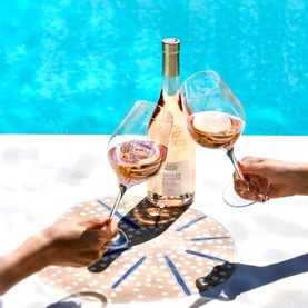 Chateau La Coste Ch&acirc;teau Ros&eacute; 75cl bottle poolside, two hands clinking filled glasses over a sunlit decorative tray